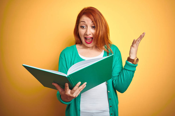 Jonge roodharige student vrouw lezen Groenboek over gele geïsoleerde achtergrond erg blij en opgewonden, winnaar uitdrukking vieren overwinning schreeuwen met grote glimlach en verhoogde handen - Foto, afbeelding