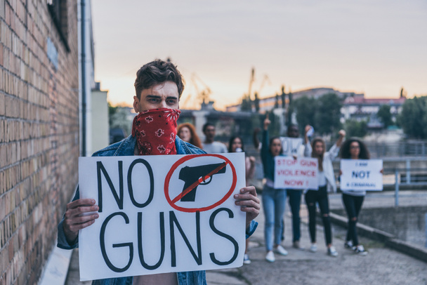 selective focus of man with scarf on face holding placard with no guns lettering near multicultural people  - Photo, Image