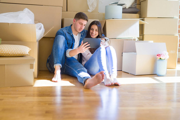 Jeune couple assis sur le sol de la nouvelle maison autour des boîtes en carton à l'aide de la tablette, souriant heureux pour le nouvel appartement
 - Photo, image