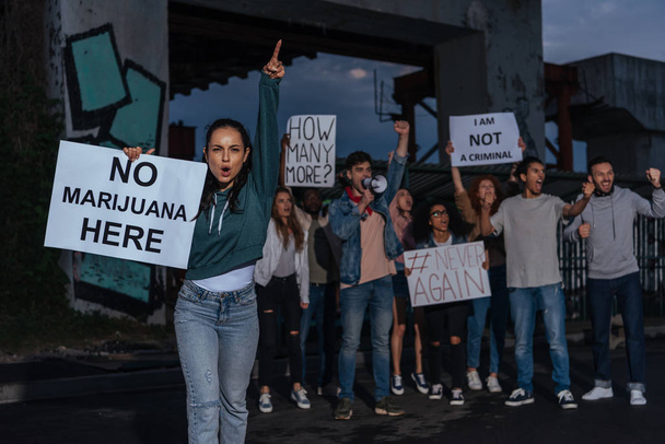 enfoque selectivo de la mujer con pancarta sin letras de marihuana cerca de personas multiculturales gritando
  - Foto, Imagen