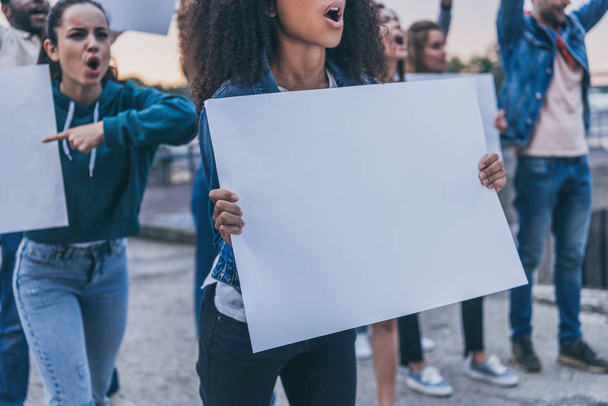 oříznutý pohled na emocionální multikulturní dívky křičící a držící během protestu prázdné cedry   - Fotografie, Obrázek