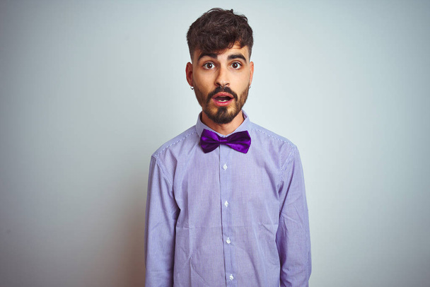 Jeune homme avec tatouage portant chemise violette et noeud papillon sur fond blanc isolé effrayé et choqué par l'expression surprise, la peur et le visage excité
. - Photo, image