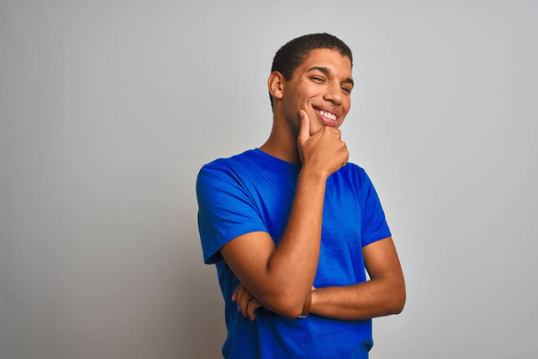 Joven hombre árabe guapo con camiseta azul de pie sobre un fondo blanco aislado mirando confiado a la cámara sonriendo con los brazos cruzados y la mano levantada en la barbilla. Pensando positivamente
. - Foto, imagen
