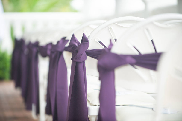 Белые свадебные кресла на острове украшены фиолетовыми луками и лентами - Фото, изображение
