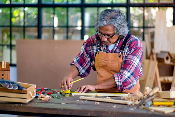 Старий майстер вимірює і працює з дерев'яними виробами, а також використовує кілька інструментів для допомоги в роботі
. - Фото, зображення