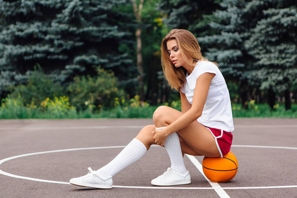 Beautiful young girl dressed in white t-shirt, shorts and sneakers, sits on a basketball court on the ball. - Foto, Bild