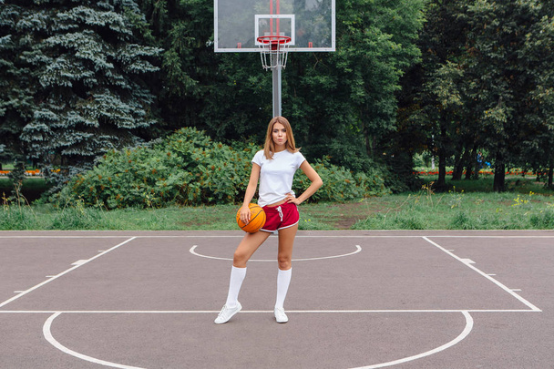 Beautiful young girl dressed in white t-shirt, shorts and sneakers, plays with a ball on a basketball court. - Фото, зображення