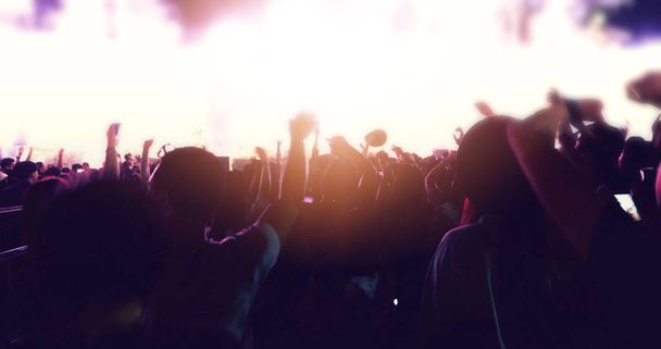 wazig van silhouetten van concert menigte op Achteraanzicht van festival menigte verhogen hun handen op heldere fase lichten - Foto, afbeelding