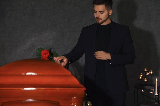 Sad young man near funeral casket with red rose in chapel - Фото, зображення