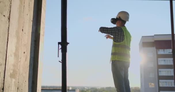Ingegnere Costruttore visualizza il piano di costruzione in piedi sul tetto dell'edificio al tramonto si erge in VR occhiali e muove le mani utilizzando l'interfaccia del futuro
.  - Filmati, video