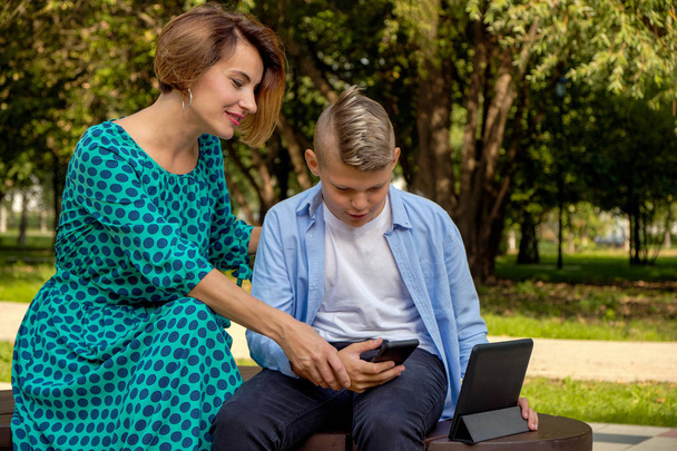 Äiti ja poika käyttävät tablettia ja älypuhelinta istuessaan luonnossa. Perhe pelaa tietokonetta tai etsii Internetistä, elektronisista laitteista ja lapsista nykyaikaisessa elämässä
. - Valokuva, kuva