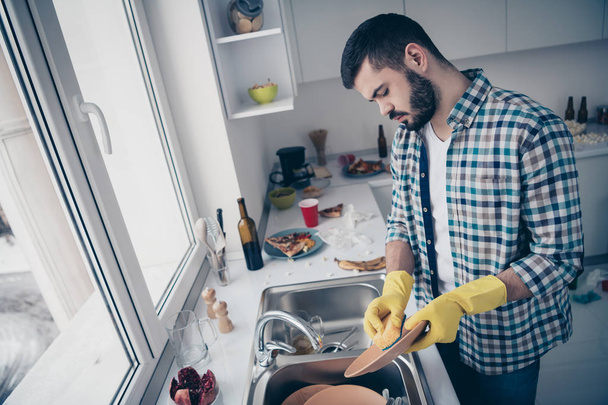 Profile side view of his he nice attractive focused hard-working guy wearing checked shirt doing domestic rub scrubbing plates in modern light white interior style kitchen indoors - Фото, изображение