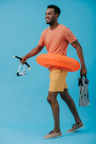 happy african american man in diving mask holding flippers and walking on blue  - Foto, Bild