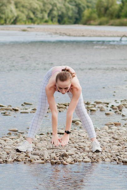 Girl in sports uniforms makes a stretch on the river bank - Photo, Image