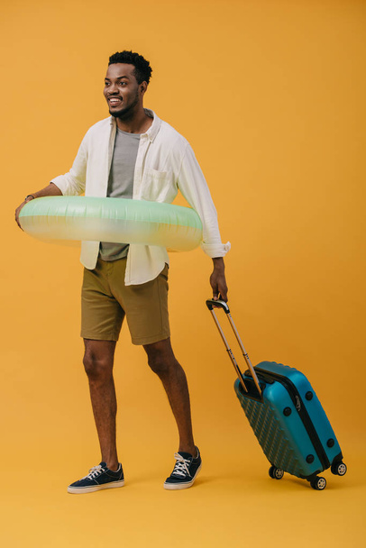 Веселый африканский американец, стоящий с плавательным кольцом и багажом на апельсине
 - Фото, изображение