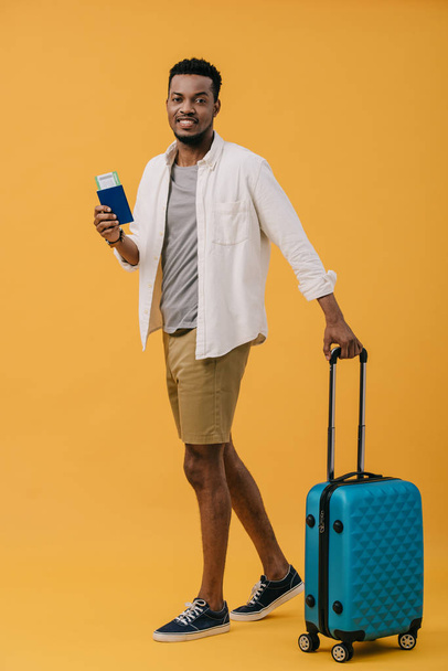 iloinen afrikkalainen amerikkalainen mies, jolla on passi lentolippuineen ja seisoo lähellä matkatavaroita oranssilla
 - Valokuva, kuva