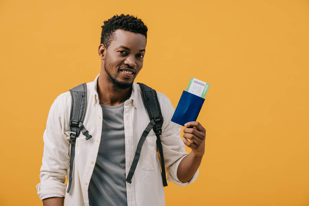 cheerful african american man with backpack holding passport with air ticket isolated on orange - Foto, Imagen