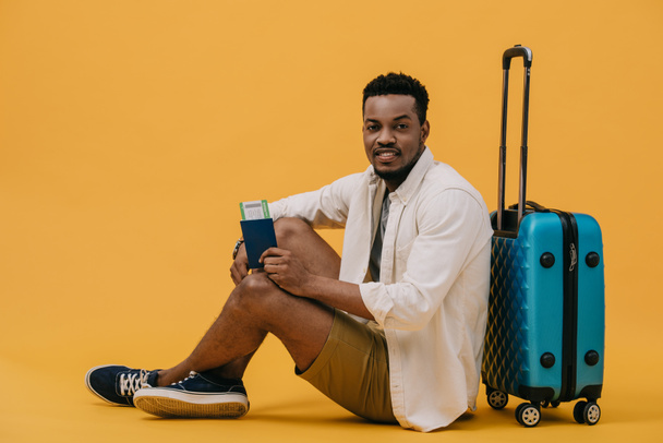 neşeli afro-amerikan adam hava bileti ile pasaport tutan ve turuncu bagaj yakınında oturan - Fotoğraf, Görsel