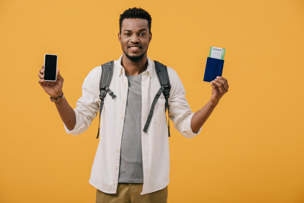 felice uomo afroamericano con zaino in possesso di passaporto con biglietto aereo e smartphone con schermo bianco isolato su arancione
 - Foto, immagini