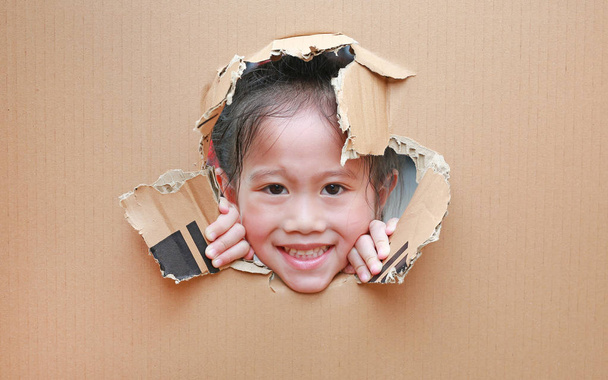 Усміхнений малюк азіатських дівчина дивиться через отвір на картоні з копіюванням простору навколо. - Фото, зображення
