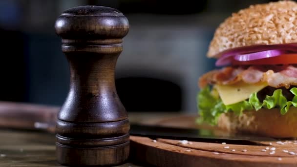 Панорамный снимок гамбургера с мягким кунжутом, жареной котлеткой, луковыми кольцами, помидорами и сыром, хрустящим беконом. На столе черные дровяные перцовые шейкеры, лежит острый нож. Кухня на заднем плане
 - Кадры, видео