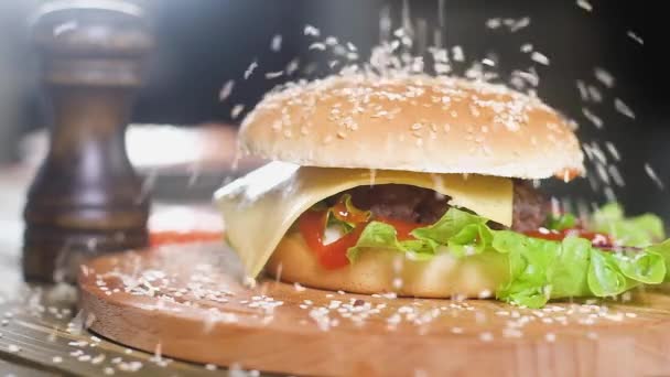 Witte sesamzaden vallen op gekookte hamburger met salade, schijfje kaas, knapperig spek en gebakken kotelet liggend op houten dienblad naast zwarte retro pepperbox. Slow Motion op de achtergrond van de keuken. - Video