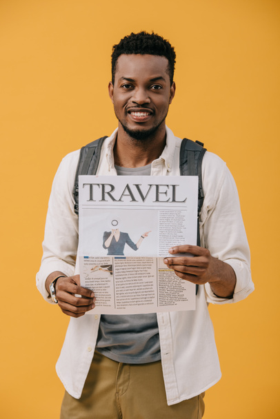 krullend Afro-Amerikaanse man staande met rugzak en het houden van reizen krant geïsoleerd op oranje - Foto, afbeelding