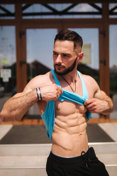 Un joven atleta entrena en el gimnasio. El culturista levanta la camisa para mostrar los músculos adbominales. Afuera.
 - Foto, imagen