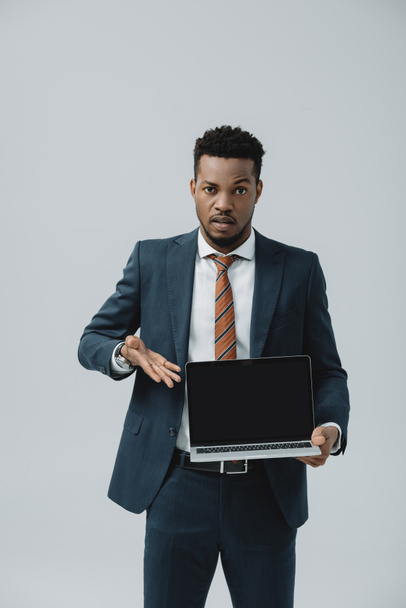 handsome african american man gesturing while holding laptop with blank screen isolated on grey  - Photo, image