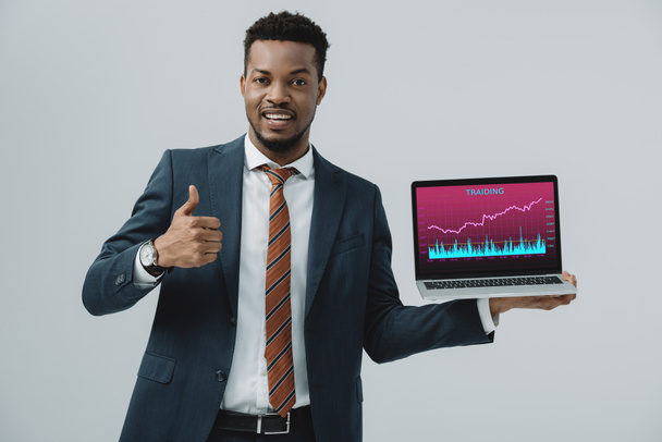 cheerful african american man holding laptop with trading analyzing  on screen and showing thumb up isolated on grey  - Foto, afbeelding