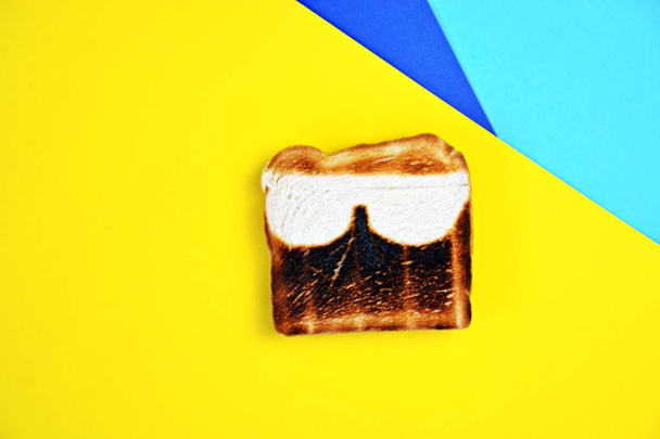 ein Toast liegt auf einer farbigen Oberfläche. der Toast wird nicht überall gleich getoastet, sondern hat ungeschmackliche Flecken in Form von Sonnenbrillen - abstraktes Konzept für Sonnenbrand im Sommer  - Foto, Bild