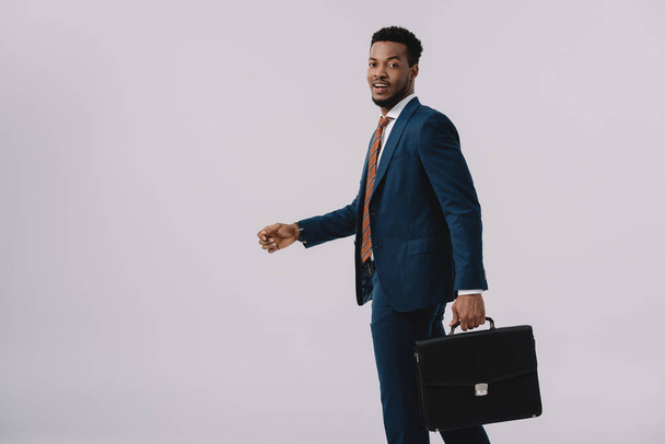 happy african american man in suit holding briefcase isolated on grey  - Foto, Bild