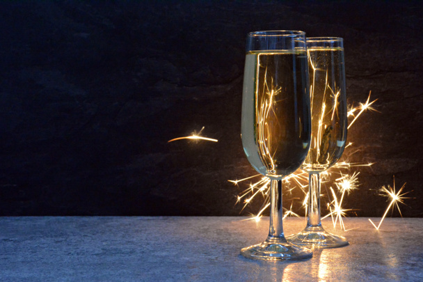 Twee gevulde Champagne fluiten staan in het donker voor een Sparkler die hen verlicht met vonken van achter-concept voor oudejaarsavond en vuurwerk met ruimte voor tekst en andere voorwerpen  - Foto, afbeelding