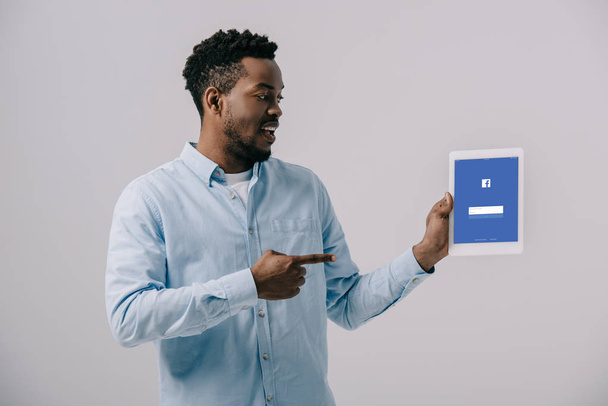 happy african american man pointing with finger at digital tablet with facebook app on screen isolated on grey  - Foto, immagini