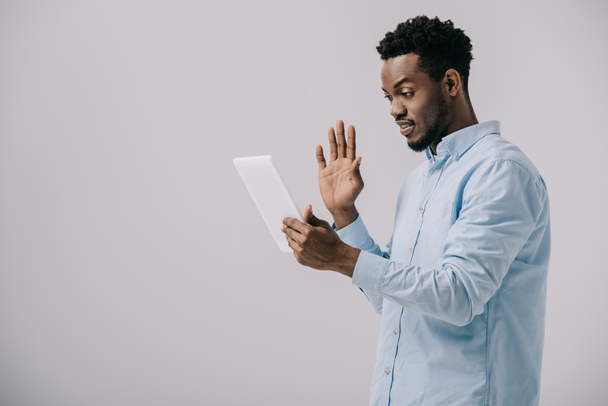 šťastný Afroameričan, který mává rukou a současně drží digitální tabletové tabletu izolovaně na šedé  - Fotografie, Obrázek
