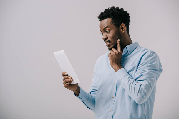 bell'uomo afro-americano che tocca il viso guardando il tablet digitale isolato sul grigio
  - Foto, immagini