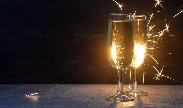 Deux flûtes à champagne remplies se tiennent dans l'obscurité devant un scintillement qui les illumine avec des étincelles par derrière concept pour la veille du Nouvel An et feux d'artifice avec espace pour le texte et d'autres objets
  - Photo, image