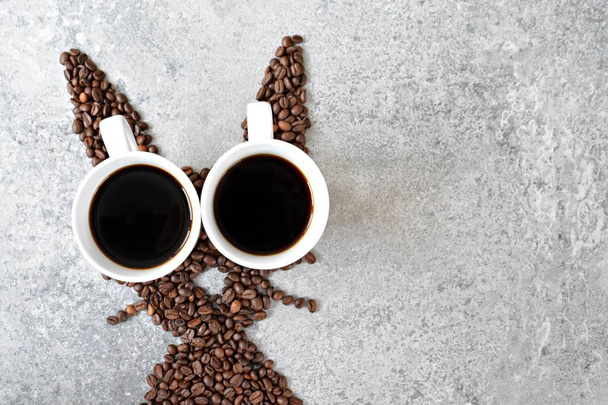 Со свежими кофейными зернами и двумя чашками свежеваренного черного кофе, заяц имеет форму на яркой мраморной поверхности - концепция с кофейными зернами в качестве подарка на Пасху - с местом для текста или других элементов
  - Фото, изображение
