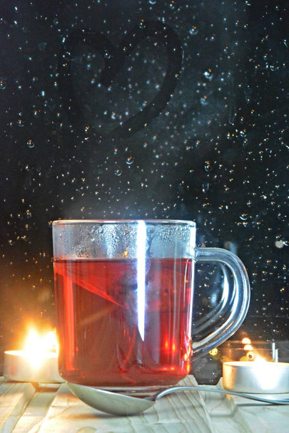 Duża szklanka herbaty owocowej stojącej na drewnianym parapecie w nocy przed deszczem-nasączone okno, w którym światła nocy można zobaczyć-przytulna deszczowa noc z filiżanką herbaty  - Zdjęcie, obraz