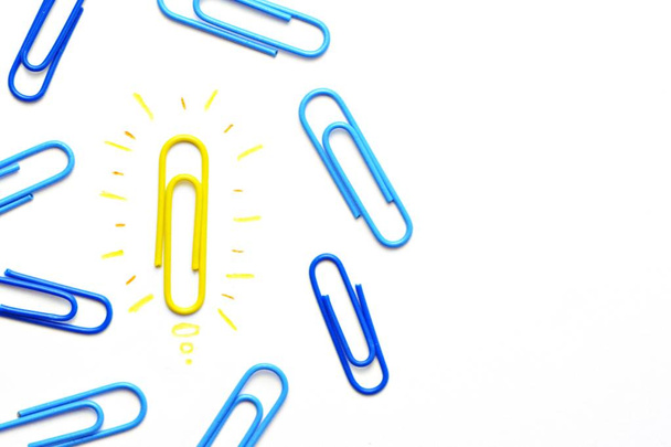 A yellow paper clip as a symbol of a light bulb that radiates lies on a white surface and symbolized the brainstorming - next to it are other paper clips without illusion  - Photo, Image