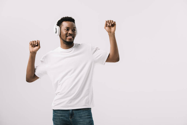pozitivní Afroameričan poslouchá hudbu ve sluchátkách a gestikuloval izolovaný na bílém  - Fotografie, Obrázek