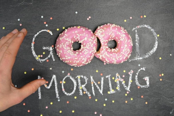 Chalk was written on a dark stone surface Good morning, the O's from the word were replaced by two pink donuts with white sprinkles - Happy start to the day with sweet donuts - Fotografie, Obrázek