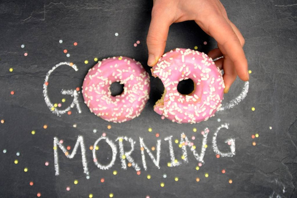 Chalk was written on a dark stone surface Good morning, the O's from the word were replaced by two pink donuts with white sprinkles - Happy start to the day with sweet donuts - Φωτογραφία, εικόνα