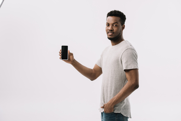 schöner afrikanisch-amerikanischer Mann steht mit der Hand in der Tasche und hält Smartphone mit leerem Bildschirm auf weißem Hintergrund  - Foto, Bild