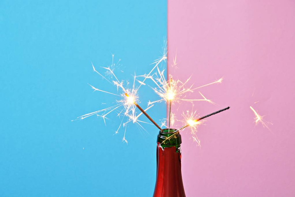 A empty champagne bottle in the neck of which a few sparklers are glowing - two toned background with a bottle of champagne in front of it and sparkles in it - Photo, Image