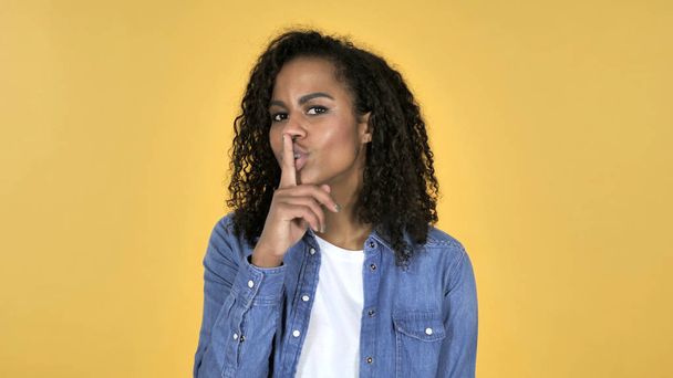 Chica africana haciendo gestos de silencio, dedo en los labios, fondo amarillo
 - Foto, imagen