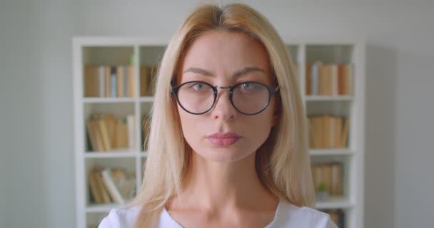 Closeup portrait of adult caucasian blonde female in glasses looking at camera indoors in the library - Filmati, video