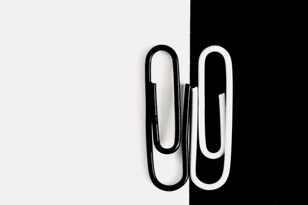 Een White-clip paperclip zit op een zwart oppervlak en een andere zwarte paperclip zit op een wit oppervlak-zwart-wit concept met paperclips-schril contrasten en verschillen als achtergrond - Foto, afbeelding