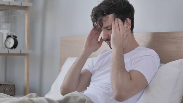 Yorgun genç Casual adam yatakta oturmuş baş ağrısı ile - Fotoğraf, Görsel