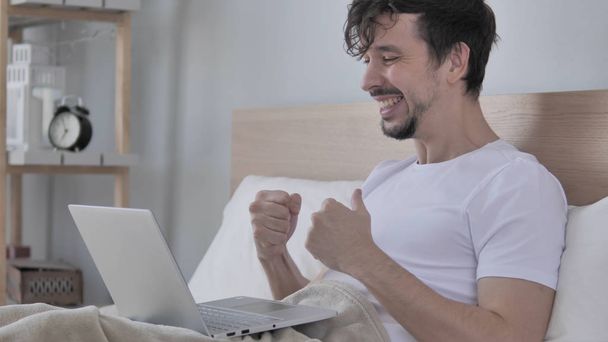 Uomo felice che celebra il successo mentre si utilizza il computer portatile a letto
 - Foto, immagini